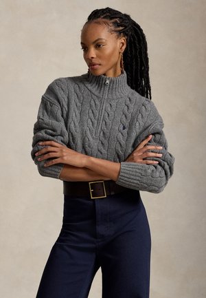 Femme aux longs cheveux tressés portant un pull gris en maille torsadée avec fermeture éclair et un pantalon sombre, bras croisés, fond neutre.