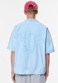 Cămașă scurtă de culoare albastru deschis, cu un design brodat pe spate ce constă din trei stele care înconjoară un cerc. Materialul pare neted, cu o croială casual.