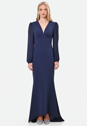 Donna che indossa un abito blu navy lungo fino a terra con maniche lunghe trasparenti, una profonda scollatura a V e tacchi alti argento, in piedi su uno sfondo semplice.