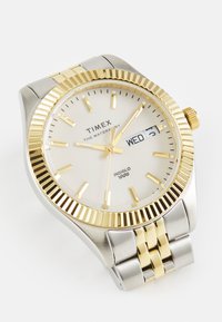 Timex ura s srebrnim nerjavečim jeklom in zlatim okvirjem. Bela številčnica vsebuje zlate kazalce, oznake ur in prikaz dneva/datuma.