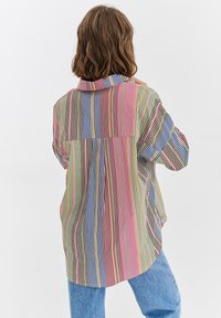 Koszula w wielokolorowe paski z krótkimi rękawami, luźnym krojem i dłuższym tylnym dołem. Materiał wydaje się lekki i gładki.