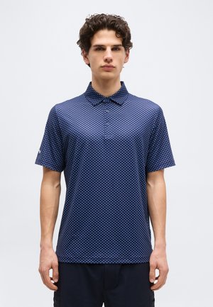 Hombre joven con cabello rizado lleva una camisa tipo polo azul marino con un pequeño patrón de diamantes blancos, de pie contra un fondo blanco liso.