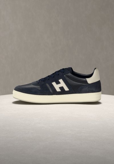 Σκούρο μπλε και μαύρο αθλητικό παπούτσι με λευκή σόλα και λεπτομέρεια στη φτέρνα, με μεγάλο λευκό "H" στο πλευρό, τοποθετημένο σε υφασμένη επιφάνεια.