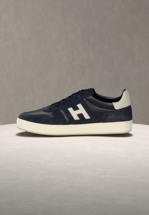 Tumši zila un melna sporta kurpe ar baltu zolīti un papēža detaļu, ar lielu baltu "H" uz sāna, izvietota uz fakturētas virsmas.