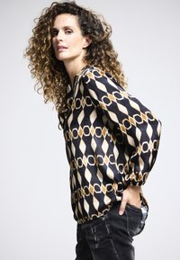 Lockere schwarze Bluse mit einem geometrischen Muster in Beige und Gold, mit einem runden Ausschnitt und Ballonärmeln, kombiniert mit ausgewaschenen schwarzen Jeans.