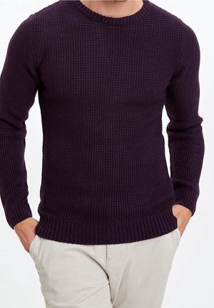 Homme portant un pull en maille texturé violet foncé avec poignets et bord côte, associé à un pantalon beige clair, mains dans les poches.