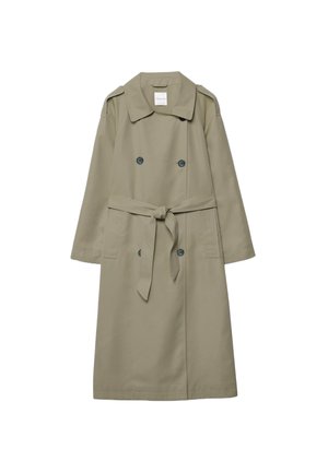 Žena oblečená v olivovom trenčkotle, hnedom pletenom topláku, bielych nohaviciach so širokými nohavicami, slamenej klobúku, slnečných okuliaroch, držiaca čiernu kabelku a čierne platformové sandále.