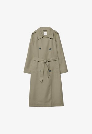 Kvinde iført olivengrøn trenchcoat, brun strikket top, hvide bukser med vide ben, stråhat, solbriller, der holder sort håndtaske og sorte platformsandaler.