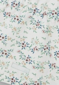Weißer Stoff mit floralem Muster, grünen Blättern, roten und blauen Blumen, kleinen gelben Akzenten und einem schlichten Etikett. Weiche Textur.