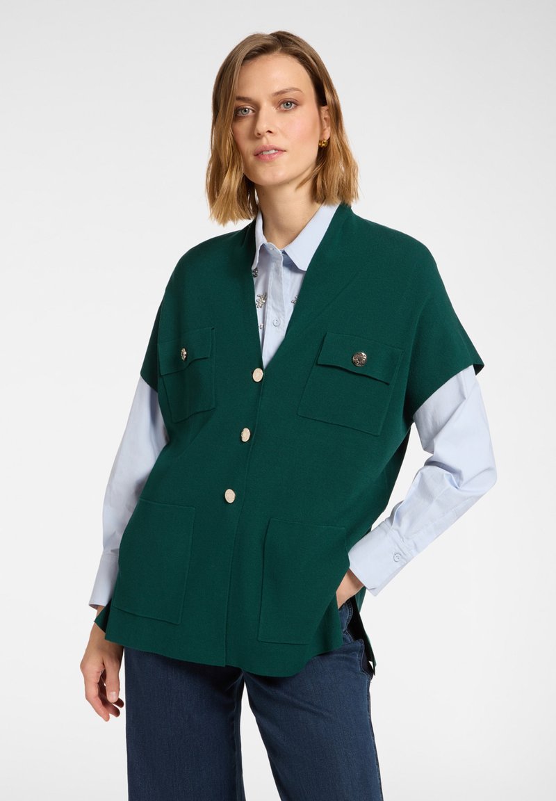 Zielona kamizelka bez rękawów z dwoma kieszeniami na piersi i zapięciem na guziki, noszona na jasnoniebieskiej koszuli. Miękka tekstura, nowoczesny design.