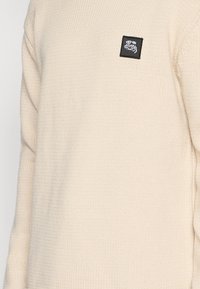 Pull en maille couleur crème avec une étiquette noire et un logo blanc sur la poitrine gauche. La texture est douce et la coupe est ample.