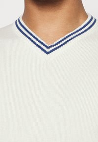 Hvid strikket sweater med V-hals med blå og hvide stribede ribkanter ved kraven. Tekstureret stofdetalje er synlig.