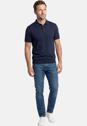 Mann steht und trägt ein marineblaues Poloshirt, blaue Jeans und weiße Sneakers vor einem einfarbigen hellen Hintergrund.