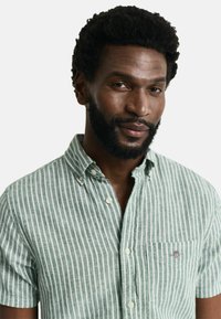 Camicetta a maniche corte a strisce verdi chiaro e bianche, con colletto abbotttonato e taschino sul petto con un piccolo emblema.