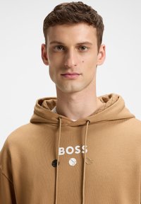 Kavos spalvos džemperis su raišteliu, su baltu "BOSS" logotipu ir trimis spalvotais teniso kamuoliukų iliustracijomis. Vidinė kapučino tekstūra su deimantiniais raštais.