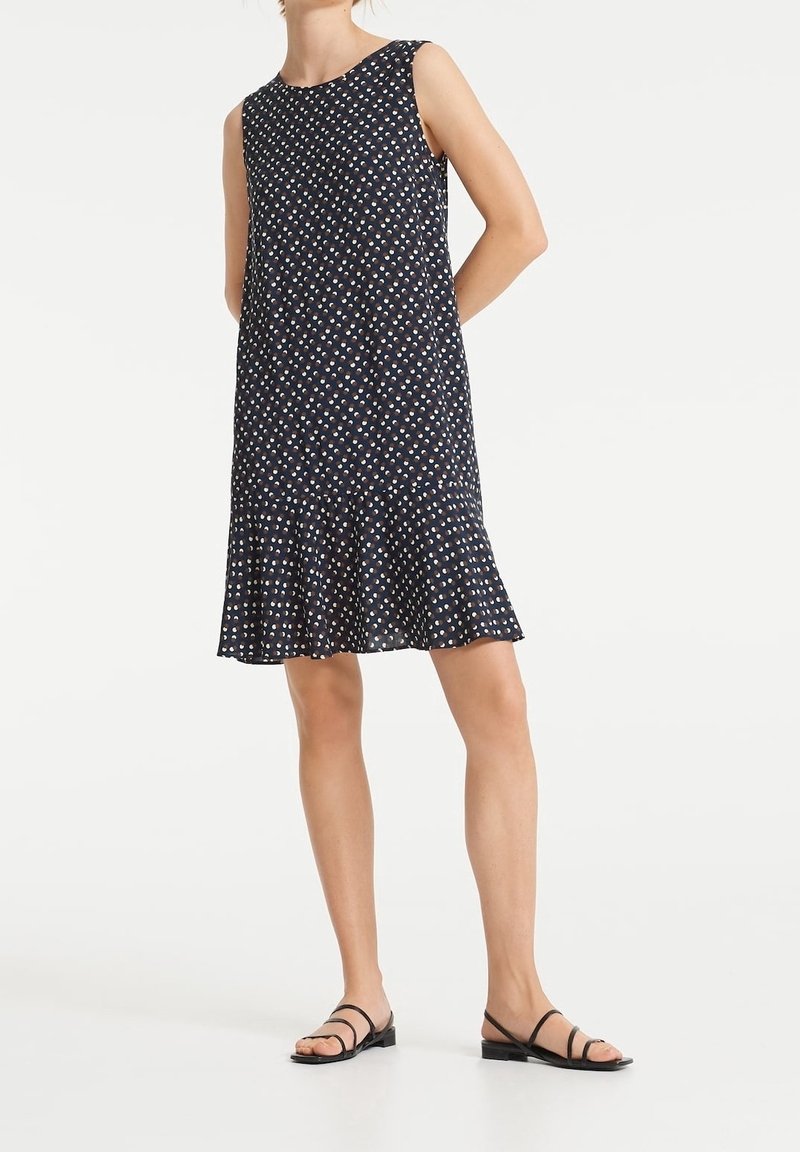 Robe sans manches bleu marine avec un petit motif floral blanc et orange, ourlet évasé, portée avec des sandales plates noires à lanières.