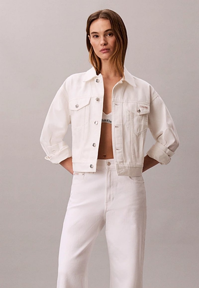 Femme aux cheveux bruns raides porte une veste en jean blanche, un pantalon blanc taille haute et un bralette blanc Calvin Klein, debout devant un fond gris.