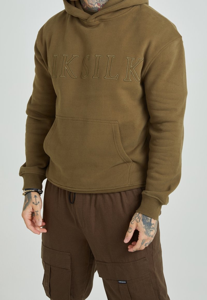 Personne portant un sweat à capuche marron avec le texte « SIKSILK » et un pantalon cargo marron assorti, les mains tatouées visibles.