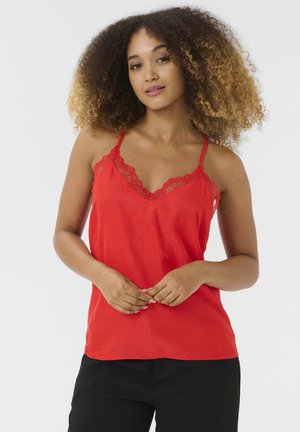 Camisola rossa con pizzo, spalline sottili e tessuto morbido. Modella che la indossa appoggiata su uno sfondo bianco semplice.
