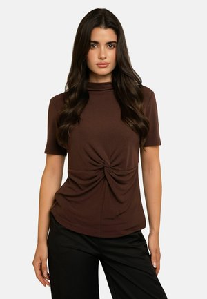 Femme aux longs cheveux foncés portant un haut marron à manches courtes avec un détail torsadé à l'avant et un pantalon noir sur un fond uni.