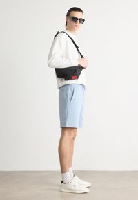Geantă de umăr neagră cu etichetă roșie, purtată peste un hanorac alb și pantaloni scurți bleu deschis. Modelul poartă ochelari de soare negri și adidași albi.