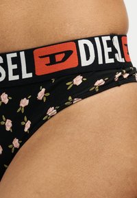 Sous-vêtements noirs à motifs floraux avec une large ceinture noire présentant le logo de la marque Diesel en blanc et rouge sur la hanche d'une personne.