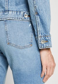 Chaqueta de mezclilla azul clara con un diseño ajustado, que cuenta con un bolsillo y botones a presión plateados, mostrando detalles desgastados y costuras visibles.