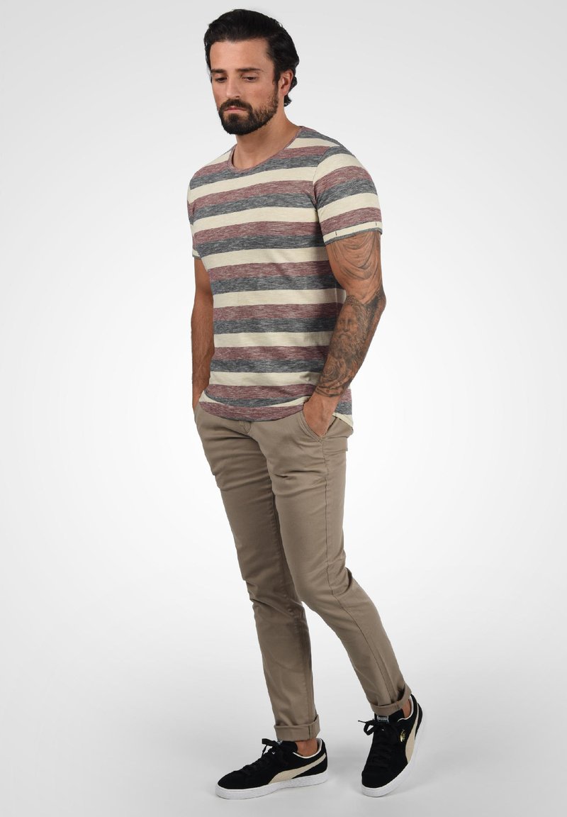 Hombre con barba y brazo tatuado lleva camiseta de rayas, pantalones beige y zapatillas negras, de pie con las manos en los bolsillos frente a un fondo blanco.