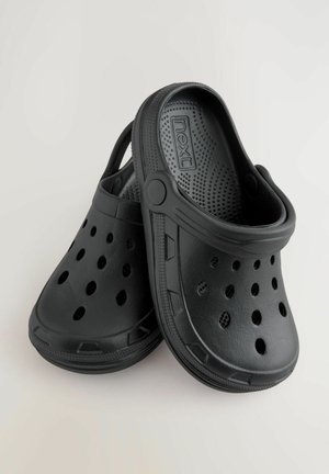 Melni plastmasas klumpes ar ventilācijas caurumiem, paceltu zoli un aizmugurējo siksniņu. Iekšpusē ir strukturēta zole, un sānos redzama preču zīme.