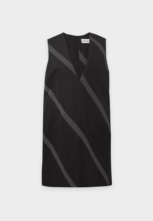 Femme aux cheveux noirs mi-longs, portant une robe noire sans manches avec des rayures grises diagonales et des chaussures plates noires, debout devant un fond uni.