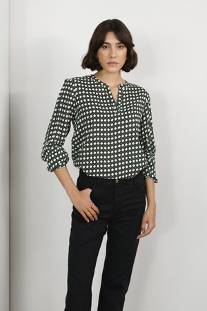 Grünes Shirt mit weißen Rautenmustern, Dreiviertelärmel und V-Ausschnitt. Kombiniert mit schwarzen Hosen. Glatte Stoffstruktur.