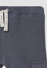 Donkergrijze katoenen leggings met een gestructureerd oppervlak, voorzien van een zijzak en een geweven label met "CLUB CUTENESS CLUB" in het wit.