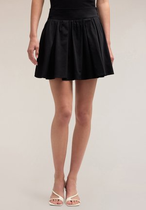 Jupe mini plissée noire avec une taille ajustée, texture en coton doux et une silhouette évasée, associée à des talons blancs à lanières.