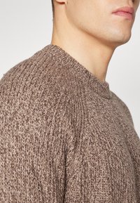 Brązowy sweter dziergany z teksturowanym wzorem, z okrągłym dekoltem i rękawami raglanowymi, prezentujący zbliżenie obszaru barku.