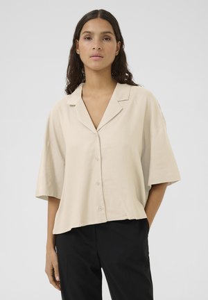 Beige short-sleeve button-up shirt with a collar, relaxed fit, and a smooth texture, paired with black trousers.