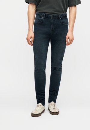 Man wearing dark slim-fit jeans and beige sneakers, standing against a plain white background.