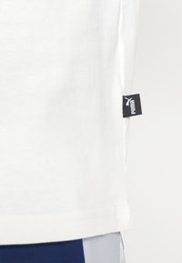 Hvid bomulds t-shirt med en lige kant, har et lille sort Puma-mærke på siden. Glat tekstur, enkel design, ingen synlige mønstre.