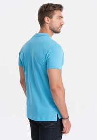 Vaaleansininen polo-paita, jossa on lyhyet hihat, sivuhalkiot helmassa ja klassinen kaulus, valmistettu pehmeästä ja hengittävästä kankaasta.