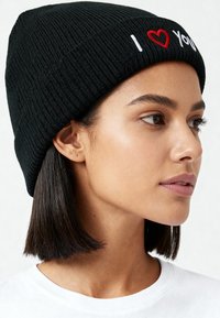 Zwarte gebreide muts met geribbelde textuur en geborduurde witte tekst "I love You" in rood en wit. Close-up zijaanzicht.