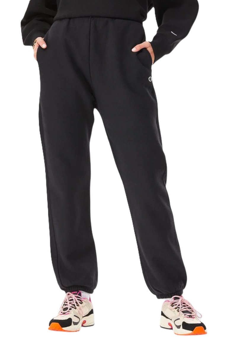 Persona vistiendo pantalones de chándal negros con bolsillos y puños elásticos, combinados con zapatillas blancas, negras y rosas con suelas naranjas.