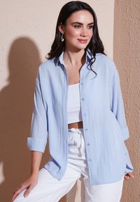 Light blue, crinkled button-up shirt with long sleeves rolled at the cuffs, paired with a white crop top and white drawstring pants.