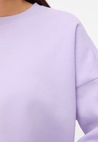 Femme portant un pull violet clair à col rond avec manches longues sur un fond uni.