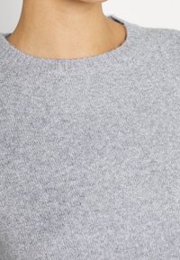 Grå stickad tröja med rund halsringning, som har en mjuk textur och ett subtilt melerat mönster. Close-up visar stickdetaljen.