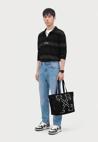 Jeune homme portant un t-shirt à manches longues rayé noir et gris, jean bleu clair, baskets blanches et noires, tenant un sac fourre-tout noir avec un motif graphique blanc.