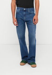 Τζιν μεσαίου μπλε σε ευθεία γραμμή, πεντατσέπη και ελαφριά φθορά. Συνδυασμένα με πράσινα αθλητικά παπούτσια και μαύρο πουκάμισο.