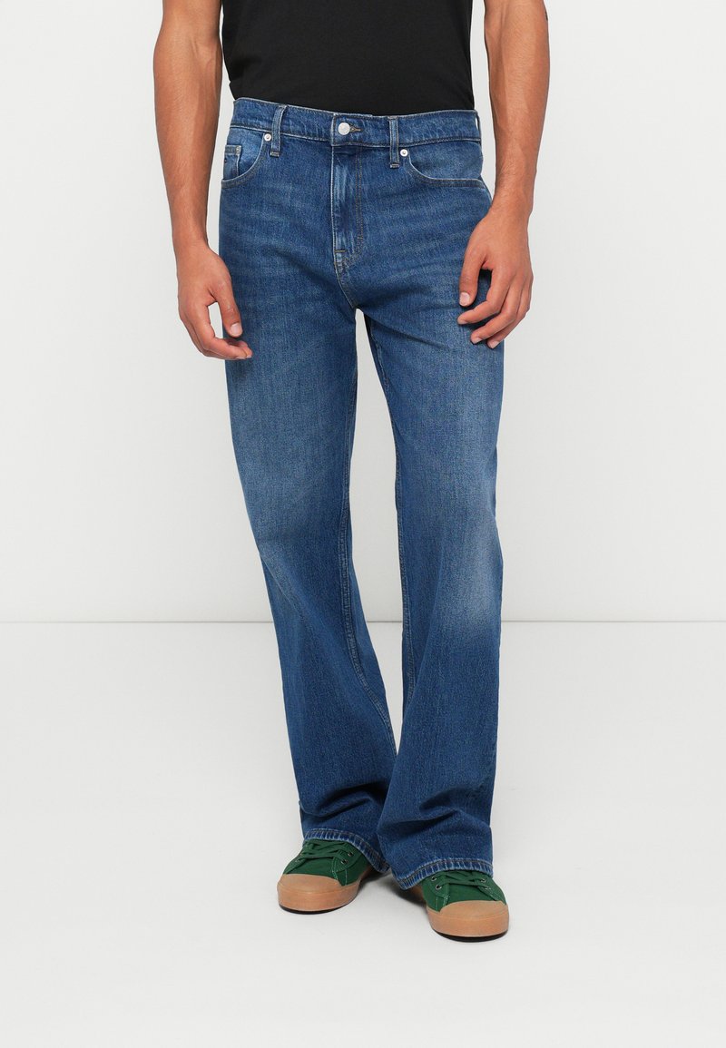 Denim jeans i mellem blå med lige ben, fem-lomme design og let falmning. Kombineret med grønne sneakers og en sort skjorte.