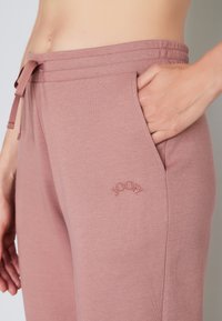 Lyserøde joggers med snor i taljen, med et broderet logo på venstre lomme og en glat, blød stoftekstur.