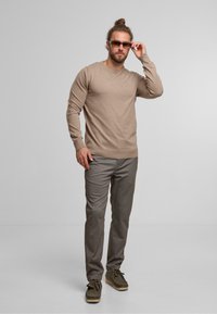 Lysebrun crew-neck sweater, mønstrede grå bukser og grønne loafers. Modellen bærer solbriller og står foran en grå baggrund.