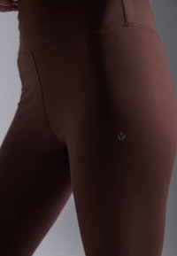 Brown leggings made of smooth, stretchy fabric, featuring a high waistband and a small logo on the thigh. Seamless design enhances comfort.