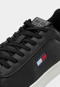 Zwarte leren sneakers met zwarte veters, witte zool en een Tommy Jeans logo patch in rood, wit en blauw aan de zijkant.